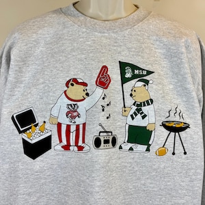May include: A gray sweatshirt featuring cartoon bears. One bear wears a red and white outfit and a foam finger, while the other holds a flag that says "MSU." The design includes a cooler, radio, grill, football, and musical notes.