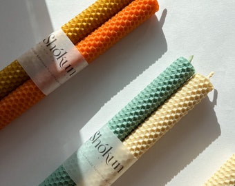 May include: Two sets of beeswax candles, one set is yellow and orange, the other is green and cream. The candles are wrapped in paper with the text "Shōkun Organics, 100% beeswax handmade" on them.