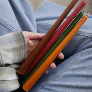 Puede incluir: Una mano sostiene cuatro velas cónicas en tonos marrón, rojo, verde y naranja. Las velas son largas y delgadas. El fondo incluye unos vaqueros azules y una camisa a cuadros.