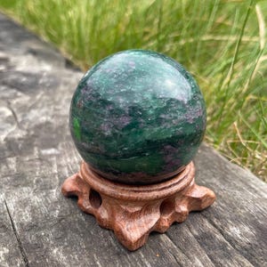 May include: A green and purple sphere, with a polished finish, rests on a carved wooden stand. The sphere has a marbled effect with green and purple swirls. The stand is a warm brown colour and has an open, decorative design. The background is blurred, showing green grass.