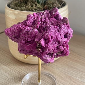 May include: A vibrant magenta, rough-textured mineral specimen displayed on a gold-toned metal stand with a clear circular base. The crystalline structure features jagged, uneven edges and deep crevices, showcasing a saturated, uniform pinkish-purple hue.