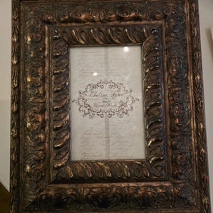 May include: A rectangular picture frame with a dark bronze finish and ornate, raised scrollwork patterns. The center insert features the text Chelsea Home 4x6 in Upscale Woods over a background of faint, cursive script writing.