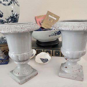 May include: Two matching, light gray urns with a distressed finish. The urns have a square base, a pedestal, and a decorative rim. The background includes a blue and white vase, a small bowl, and other decorative items.