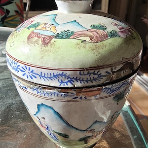 May include: A vintage, hand-painted ceramic jar with a lid. The jar features a detailed landscape scene with figures, mountains, and trees in shades of blue, green, and brown. The rim has a blue floral pattern. The jar is approximately 8 inches tall.