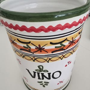 May include: A white ceramic wine cooler with a green rim and base. The cooler features a decorative design with red, yellow, orange, and black accents. The word "VINO" is printed on the front in black.