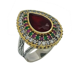 May include: A silver ring with a large, teardrop-shaped, faceted, dark red gemstone at the center. The gemstone is surrounded by a gold-colored setting with small white, green, and red stones. The ring has a textured silver band.