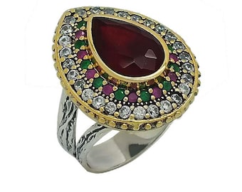 May include: A teardrop-shaped ring featuring a large central dark red stone surrounded by concentric circles of small green, pink, and clear stones. The setting is gold-toned, while the band has a textured, oxidised silver-coloured finish.
