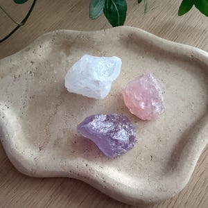 Op de afbeelding: Een beige, organisch gevormde schaal bevat drie ruwe kristallen: een heldere kwarts, een roze rozenkwarts en een paarse amethist. De kristallen zijn ruw bewerkt en variëren in grootte. De schaal staat op een houten oppervlak, met groene bladeren op de achtergrond.