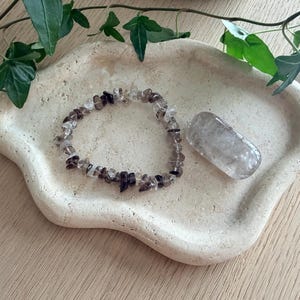 Op de afbeelding: Een armband gemaakt van kleine, onregelmatig gevormde rookkwarts kralen. Naast de armband ligt een gladde, ovaalvormige rookkwarts steen. Beide items liggen op een lichtbeige, organisch gevormd dienblad, met groen blad in de achtergrond.
