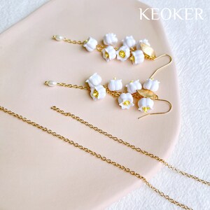K&ouml;nnte beinhalten: Ein Paar goldene Kettenohrringe mit wei&szlig;en Maigl&ouml;ckchen-Bl&uuml;tenanh&auml;ngern. Die Ohrringe werden auf einer rosa Oberfl&auml;che mit dem Wort "KEOKER" in wei&szlig;er Schrift pr&auml;sentiert.