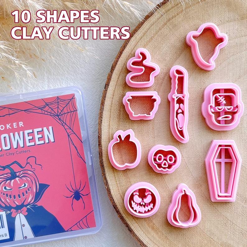 May include: A set of ten pink Halloween-themed clay cutters in various shapes, including a ghost, cauldron, knife, and pumpkin. The cutters are displayed on a wooden surface next to a pink box with Halloween-themed graphics and the text "10 SHAPES CLAY CUTTERS".