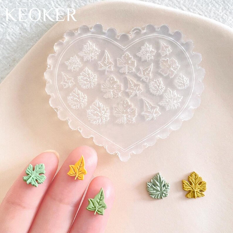 May include: A heart-shaped silicone mold with 16 leaf designs. The mold is clear and has a scalloped edge. There are four small, colorful leaf-shaped decorations on a white surface.