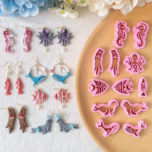 Puede incluir: Una colección de pendientes hechos a mano con criaturas marinas, como caballitos de mar, pulpos, ballenas, peces y focas. Los pendientes son de varios colores, como rosa, azul y marrón. También se incluyen cortadores de arcilla rosa con forma de criaturas marinas.