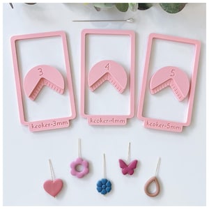 May include: Pink polymer clay earring making tools and earring designs. The tools include three pink rectangular frames with semi-circular cutouts labeled 3, 4, and 5. Below are earring designs in the shape of a heart, flower, blue flower, butterfly, and teardrop.