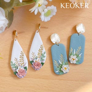 Op de afbeelding: Twee paar bloemenoorbellen. Het ene paar is wit met roze en witte bloemen en gouden accenten. Het andere paar is blauw met witte bloemen en gouden accenten. De oorbellen zijn handgemaakt en hebben een delicate, vrouwelijke uitstraling.