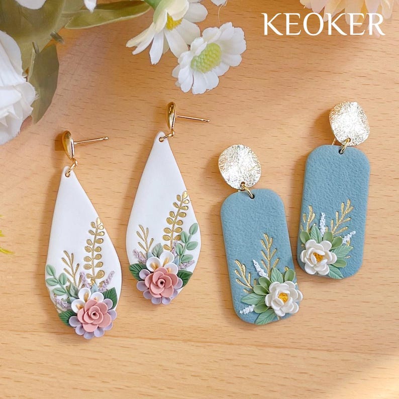 Op de afbeelding: Twee paar bloemenoorbellen. Het ene paar is wit met roze en witte bloemen en gouden accenten. Het andere paar is blauw met witte bloemen en gouden accenten. De oorbellen zijn handgemaakt en hebben een delicate, vrouwelijke uitstraling.