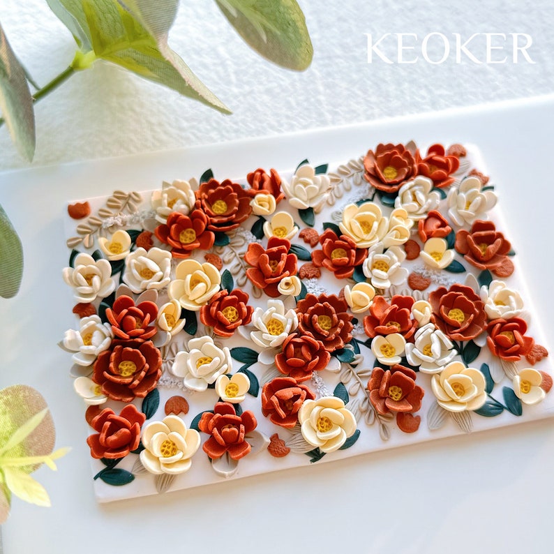 May include: A close-up of a handmade floral arrangement with orange and cream flowers. The flowers are made of polymer clay and are arranged on a white background. The text "KEOKER" is visible in the upper right corner of the image.