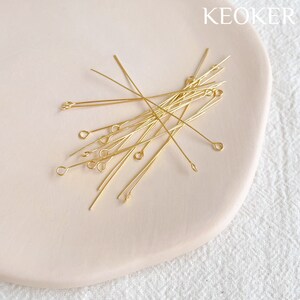 Pu&ograve; includere: Primo piano di un mucchio di spilli per la testa dorati con un anello a un'estremit&agrave;. Gli spilli sono disposti su una superficie rosa chiaro.