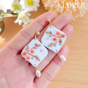 May include: A pair of white square earrings with delicate pink and green floral designs and a pearl dangle. The earrings are held in a hand.