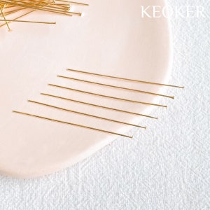 May include: A close-up of a group of gold head pins on a pink surface. The pins are arranged in a row, with the heads facing the viewer.