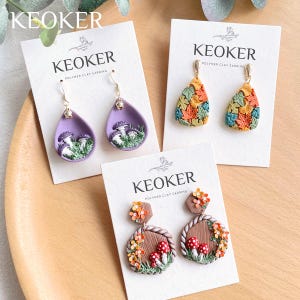 May include: Three pairs of polymer clay earrings with autumnal themes. The first pair features purple teardrop-shaped earrings with white mushrooms and green foliage. The second pair features teardrop-shaped earrings with orange, yellow, and green leaves. The third pair features brown, round earrings with red mushrooms, orange flowers, and green foliage. All earrings are labeled "KEOKER POLYMER CLAY EARRING".