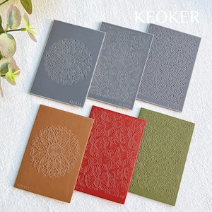 May include: A selection of embossed notebooks in shades of grey, brown, red, and green. Each notebook displays a unique floral or mandala pattern. The brand name "KEOKER" is printed at the base of each notebook.