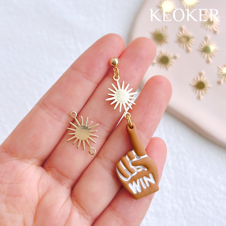 May include: Gold and brown dangle earrings featuring a sun and a hand with the word "WIN" on the finger. The earrings are shown on a hand.