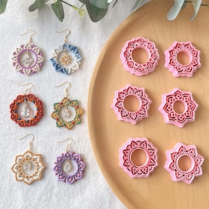 May include: A selection of colourful, handcrafted earrings with intricate floral designs and crystal accents. The earrings are displayed alongside pink, flower-shaped moulds on a wooden tray, showcasing a variety of styles and colours.