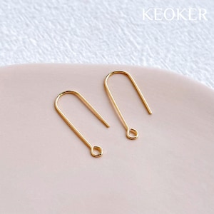 May include: Two gold-toned earring hooks, each with a small loop at the end. The hooks are on a light pink surface.