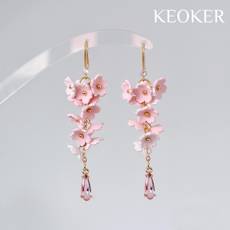 May include: A pair of gold-toned earrings featuring cascading pink flower clusters and teardrop-shaped pink gems. The earrings have a delicate design with small clear crystals and are set against a soft, neutral background.