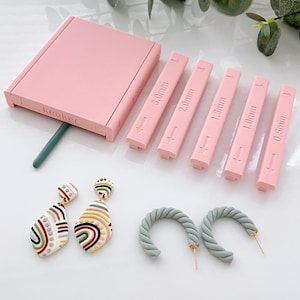 May include: A pink clay tool kit with the word "keaker" on the side, and six pink measuring tools labeled 0.5mm to 3.0mm. Two pairs of earrings are also shown: one pair with a teardrop shape and colorful patterns, and another pair of gray twisted hoop earrings.