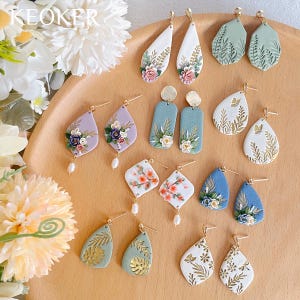 May include: A collection of 12 pairs of handmade polymer clay earrings. The earrings feature various designs, including floral patterns, leaves, butterflies, and geometric shapes. The earrings are all made with gold-toned metal findings.