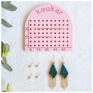 Peut inclure: Outil de fabrication de boucles d'oreilles rose avec une règle graduée en centimètres, deux paires de boucles d'oreilles vertes et dorées et quatre tiges de boucles d'oreilles dorées.