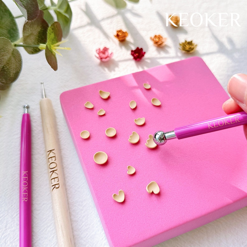 May include: A pink silicone mat with various shapes for crafting polymer clay. The mat has a light brown polymer clay in the shapes. A person is using a pink tool with a silver ball on the end to smooth the clay.