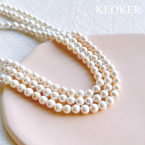 Peut inclure: Un collier de perles blanches à trois rangs. Le collier est posé sur une surface rose.