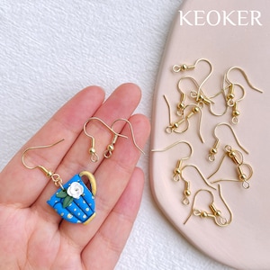 May include: A set of gold earring hooks with a blue and gold teapot charm earring. The charm features a white flower and gold polka dots. The text "KEOKER" is visible in the background.