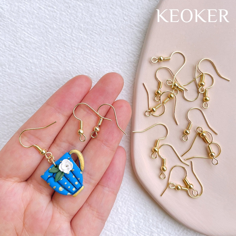 May include: A set of gold earring hooks with a blue and gold teapot charm earring. The charm features a white flower and gold polka dots. The text "KEOKER" is visible in the background.