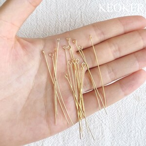 Pu&ograve; includere: Primo piano di una mano che tiene un gruppo di spilli per la testa dorati. Gli spilli per la testa misurano circa 3,8 cm di lunghezza e hanno un piccolo anello nella parte superiore.