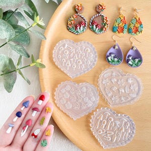 Könnte beinhalten: Vier herzförmige Silikonformen zum Herstellen von Polymer Clay Ohrringen. Die Formen zeigen verschiedene Designs, darunter Pilze, Blätter und Bäume. Die Formen sind auf einer Holzoberfläche mit Beispielen der fertigen Ohrringe abgebildet.