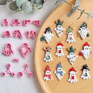 May include: A collection of pink plastic clay cutters in various shapes alongside a set of white ghost-shaped earrings decorated with holiday themes like Santa hats, scarves, reindeer antlers, and Christmas lights, all featuring gold-toned earring hooks.