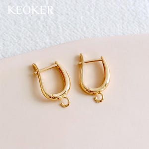 May include: Two gold-colored hoop earrings with a curved design and a small loop at the bottom for attaching charms. The earrings are displayed on a light pink surface. The word "KEOKER" is visible in the top left corner.