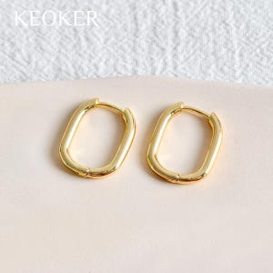 Op de afbeelding: Een paar goudkleurige, ovaalvormige oorringen. De oorbellen hebben een gladde, gepolijste afwerking en een eenvoudig ontwerp. De merknaam "KEOKER" is linksboven zichtbaar.