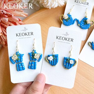 May include: Two pairs of blue and gold teapot and teacup earrings with white flowers. The earrings are on white cards with the text "KEOKER - POLYMER CLAY EARRING -" printed on them.