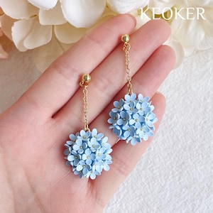 Puede incluir: Un par de pendientes delicados con racimos de flores azul claro. Cada pendiente tiene una cadena dorada y una pequeña bola dorada en la parte superior. Los pendientes son hechos a mano y tienen un tema floral.