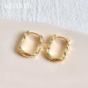 May include: A pair of gold-colored rectangular hoop earrings. The earrings have a textured, hammered appearance. The word "KEOKER" is printed in white in the upper left corner. The earrings are displayed on a light pink surface.