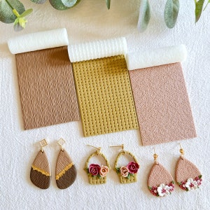 Op de afbeelding: Een set van zes polymer clay oorbellen met bloemen- en mandontwerpen. De oorbellen worden getoond op drie verschillende textuur oppervlakken: bruin, goud en roze.