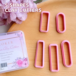 Può includere: Cinque tagliabiscotti in plastica rosa di diverse forme, tra cui un rettangolo, un ovale e un rettangolo lungo. I tagliabiscotti sono su una superficie di legno. Il testo "5 SHAPES CLAY CUTTERS" è in alto nell'immagine.