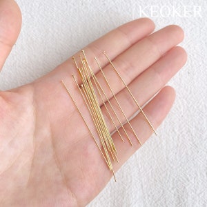 May include: A close-up of a hand holding a pile of gold-colored head pins. The pins are about 2 inches long and have a rounded head.