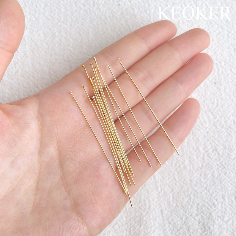 May include: A close-up of a hand holding a pile of gold-colored head pins. The pins are about 2 inches long and have a rounded head.