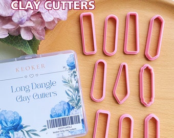 May include: Ten pink plastic clay cutters in various long shapes, including rectangles, ovals, and a pointed shape. The cutters are arranged on a wooden surface with a floral patterned box in the background. The box has the text "KEOKER Long Dangle Clay Cutters" and a barcode with the text "X003TUMQLZ keoker long clay cutters".
